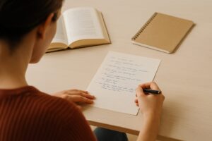 Person schreibt handschriftlich Dialoge auf einem hellen Holztisch, daneben ein geöffnetes Buch und ein Notizbuch – symbolisch für kreatives Schreiben.